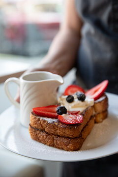 waitress with dark apron holding plate of french toast with fresh berries and cream with a syrup pitcher on a white plate