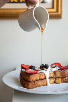 hand pouring maple syrup from white ceramic jar onto french toast with fresh berries and cream on a white plate in modern minimal cafe