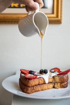 hand pouring maple syrup from white ceramic jar onto french toast with fresh berries and cream on a white plate in modern minimal cafe