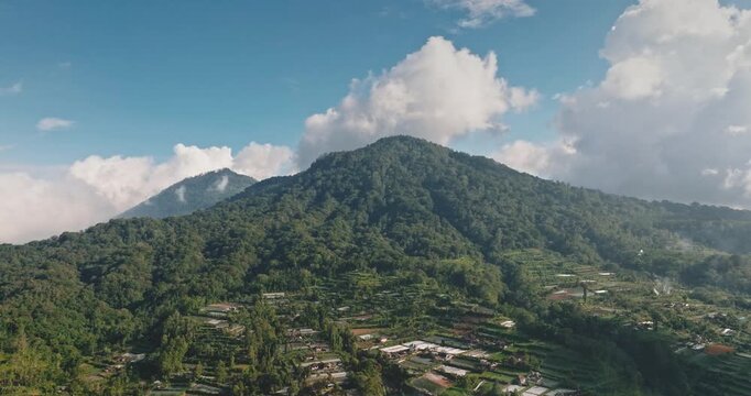 Lush green mountains peak covered in tropical forest and terraced farmlands dominate the rural landscape of traditional small village under blue sky. Explore Bali, Indonesia. Aerial zoom out panorama