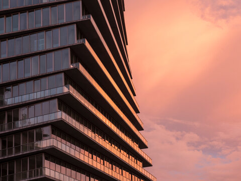 Close-up view of a contemporary residential high-rise at sunset, with stacked cantilevered balconies and floor-to-ceiling glass windows bathed in warm golden-pink light. 