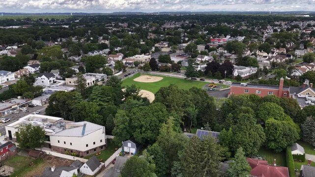 A baseball diamond borders buildings and West Hempstead neighborhoods, Nassau County on Long Island, New York. The drone travels northeast at nearly level altitude. Trees and buildings share the view.