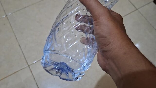 Hand holding empty plastic mineral water bottle with a small remaining amount of water inside over tile floor background