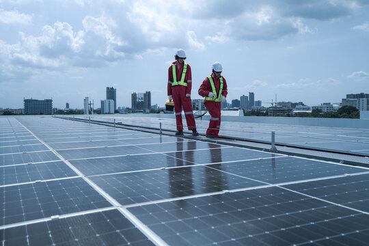 Two solar technicians in red safety suits walk a vast photovoltaic rooftop carrying out checks overlooking the city, showing solar power generation, inspection and maintenance on a rooftop.