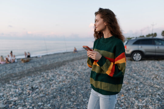 Young woman in a green striped sweater holds a smartphone while standing on a pebble beach at sunset. Blurred people and a car in the background. Concept of travel and leisure.