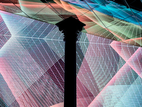A dark silhouetted classical column with an ornate capital stands against a vivid backdrop of intersecting colorful lines and geometric grid patterns.