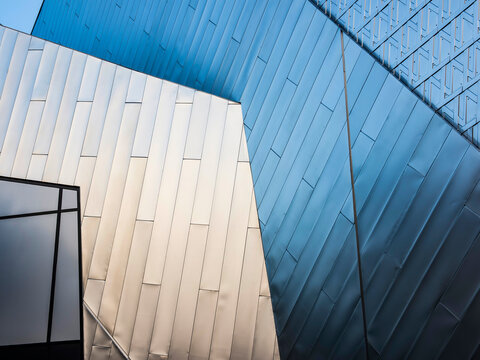 Abstract close-up of intersecting angular architectural surfaces in blue and white metallic panels, emphasizing geometric form, texture, light, and shadow.