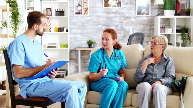 Two nurses consult with a senior woman as one records notes on a clipboard, illustrating coordinated elder care, communication and care planning.