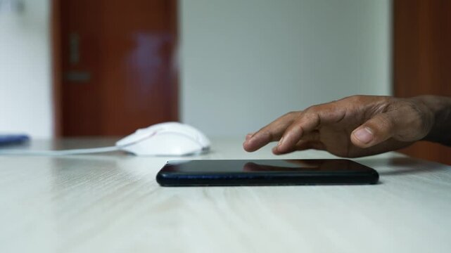Hand Using Smartphone on Home Office Desk, Close-up of a person's hand using a smartphone on a light wooden table, with a computer mouse and blurred home office background.