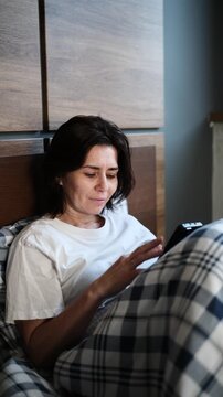 Woman relaxes in bedroom, checking smartphone under checkered blanket