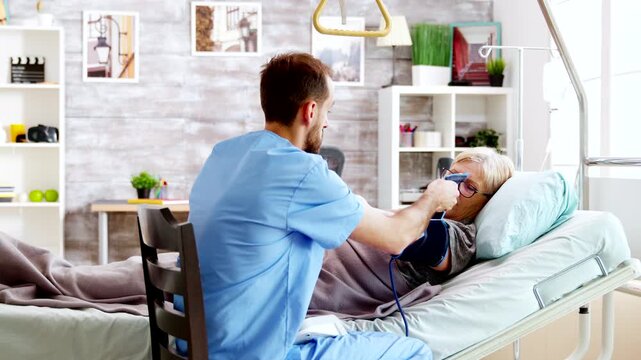 Male nurse measures the blood pressure of an older patient resting in bed, supporting routine vital-sign monitoring and professional senior healthcare.