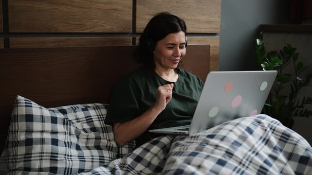Woman sitting in bed with laptop, smiling and listening to music in relaxed home setting