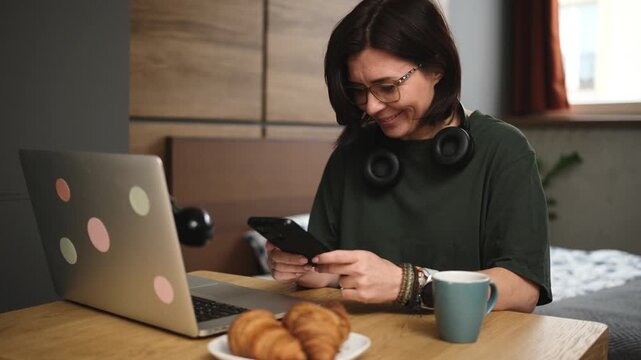 Woman taps her smartphone with both hands at the kitchen table, enjoying coffee and a croissant