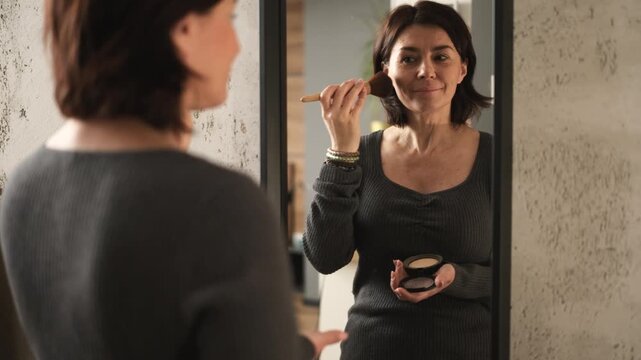 Woman applies makeup in indoor setting with gray sweater visible in front of mirror glass