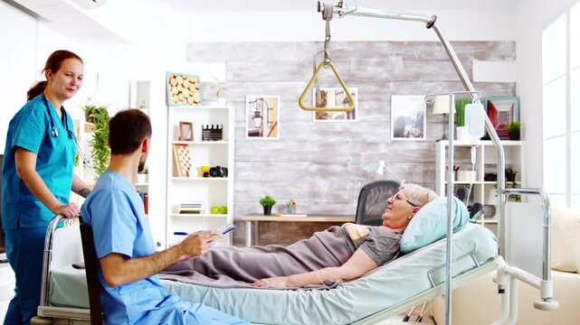 Two nurses consult an older patient in bed while one takes notes, showing coordinated care, clinical teamwork, communication and senior health support.