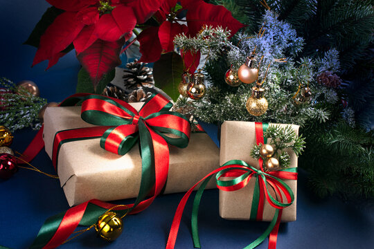 Christmas decor with handmade gift boxes with bow, red poinsettia, decorative fir branch, bells and balls for tree decoraton on blue background. New Year winter holiday composition with presents.