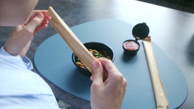 Unrecognizable person unpacking disposable wooden chopsticks to eat delicious asian yuba bowl with vegetables from takeout container, quick healthy lunch.
