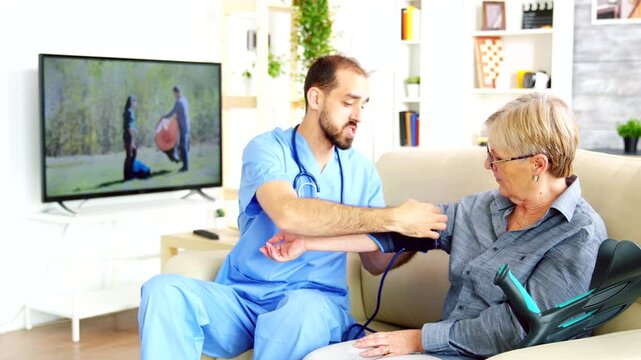 Male healthcare worker measures blood pressure of a senior woman seated on a sofa, combining routine monitoring, preventive care and senior support, also emphasizing checkup and measurement.