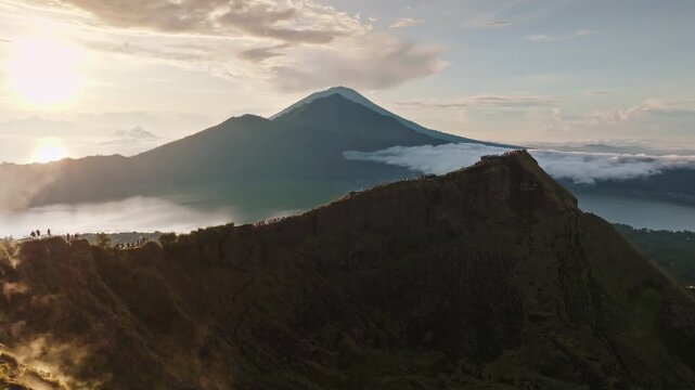 Hikers people relax on Mount Batur crater rim watch golden sunrise over misty caldera and lake, with Mount Agung rising in the distance serene Bali volcanic panorama at dawn. Aerial view drone flight