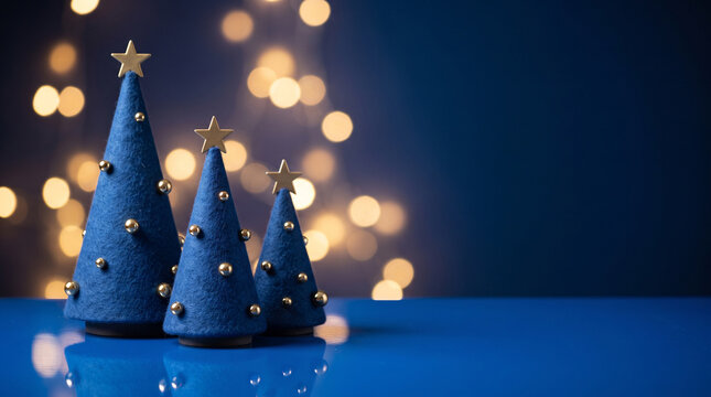 Three decorative blue felt christmas trees with gold stars and ornaments on a reflective blue surface with soft bokeh lights in the background