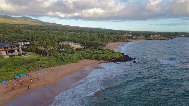 Aerial view of golden sand beach on Maui Hawaii island USA with swimmers surfers and sunshades palm trees luxury homes green hills and turquoise ocean waves rolling in at sunset light.