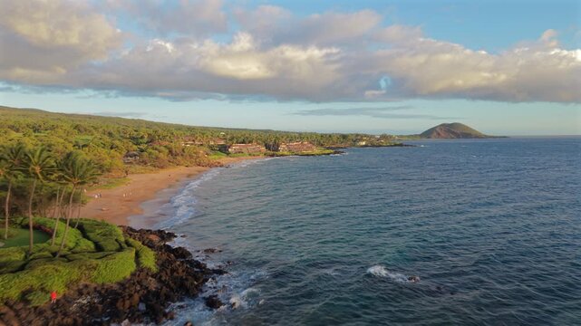 Aerial view of Maui Hawaii island USA at golden hour with turquoise surf rolling onto sandy beach palm trees lush green coastline resort homes and volcanic cinder cone under bright clouds.