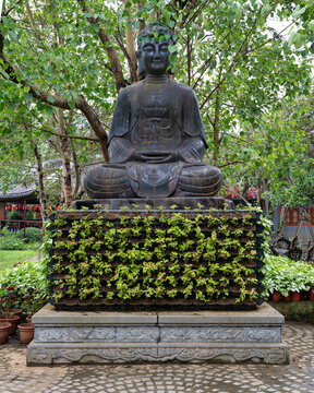 Black Buddha statue on a pedestal with vertical planters and green plants under a tree at the Bai Dinh Pagoda in Ninh Binh, Vietnam