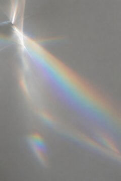 A soft, dreamy abstract photograph featuring prismatic rainbow light and subtle spectral reflections across a muted gray surface.