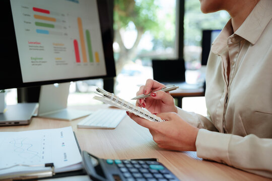 Asian businesswoman analyzing financial data and business charts while taking notes and reviewing reports at a modern office desk.