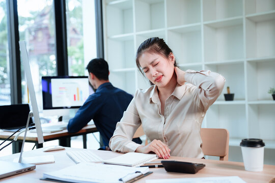 Asian businesswoman experiencing back pain while working at a desk in a modern office, with a colleague working at a computer in the background.