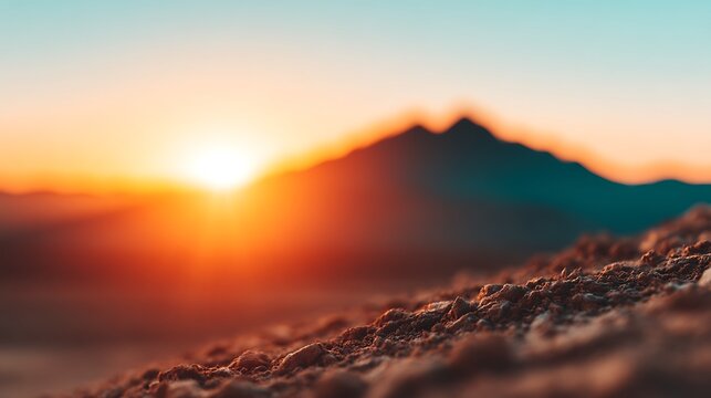 A vibrant sunrise illuminates a mountain silhouette with warm orange and blue tones, highlighting textured rocky foreground in a serene natural landscape.
