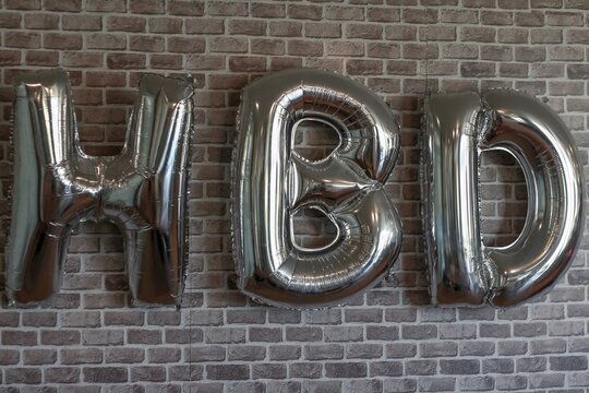 Silver HBD foil letter balloons hanging on brick wall background. 