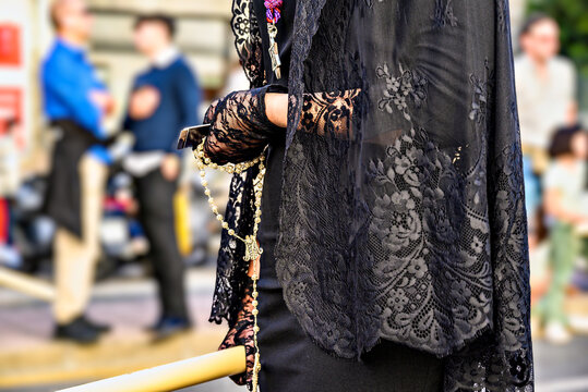 A woman dressed in traditional manola attire (with a black mantilla), holding a rosary and a large candle during a Holy Week procession in Granada, Andalusia, Spain.