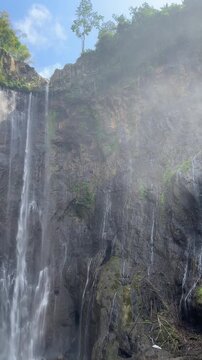 Vertical shot looking up at thin waterfall streams trickling down a misty cliff face topped with tropical vegetation in East Java, Indonesia
