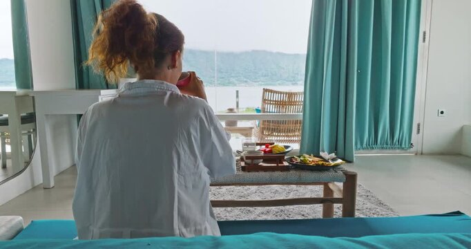 Woman drinking coffee and enjoying a healthy breakfast of tropical fruits and omelette overlooking lake Batur in balcony window, embracing a serene morning. Travel and relax on Bali resort. Back view