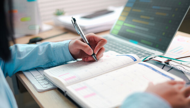Close up woman hand writing schedule event agenda on calendar notebook with pen. Businesswoman planning work task project online on laptop desk at home office.