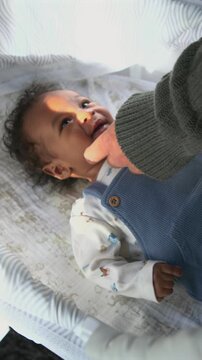 Vertical Screen: African American infant boy smiles and coos while lying on soft patterned blanket in bassinet as parent hand gently touches his cheek in warm morning light indoors.