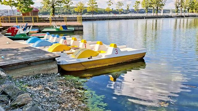 Colorful pedal boats are moored along a wooden pier on a calm lake, with reflections of the sky and surrounding waterfront creating a peaceful summer leisure scene