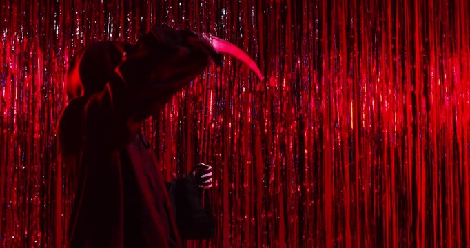 Male funny person in Halloween death costume and skull mask having fun and dancing on festive red foil background holding scythe celebrating holiday. Halloween party and horror concept. 4k video.