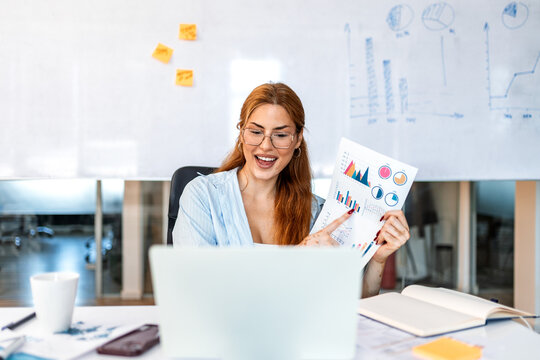 Business woman presenting charts on video call. Female professional online meeting, corporate presentation, remote communication. Smiling manager holding financial report and pointing at laptop.