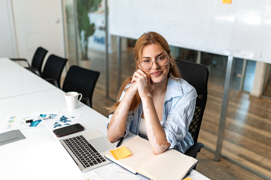 Business woman sitting at desk in modern office. Smiling female professional looking at camera, resting chin on hands. Workplace interior with laptop computer, papers, coffee cup, and conference room.