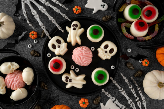 Halloween jelly desserts shaped like spooky ghosts, skulls and eyeballs, served on black plates with pumpkins and festive decorations. Top view, copy space.