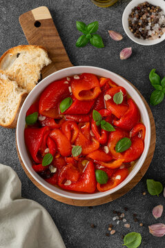Roasted red peppers in olive oil with garlic and basil in a bowl on dark background. Mediterranean appetizer served with bread. Top view.