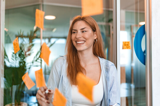 Business woman writing on sticky notes on glass wall in modern office. Smiling female professional brainstorming strategy, agile project management, planning workflow, creative startup concept.