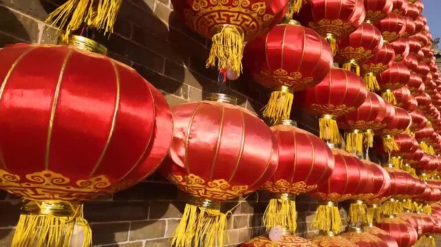 Traditional red Chinese lanterns with gold tassels and auspicious patterns hanging in rows on an aged brick wall under sunlight