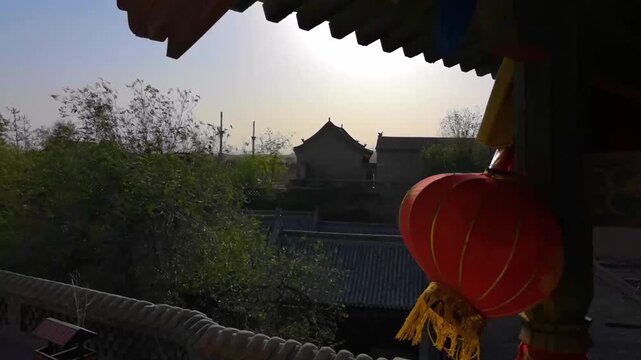 Red Chinese lantern with gold tassels hangs from ornate eaves, framing a sunlit traditional courtyard with tiled rooftops and green trees