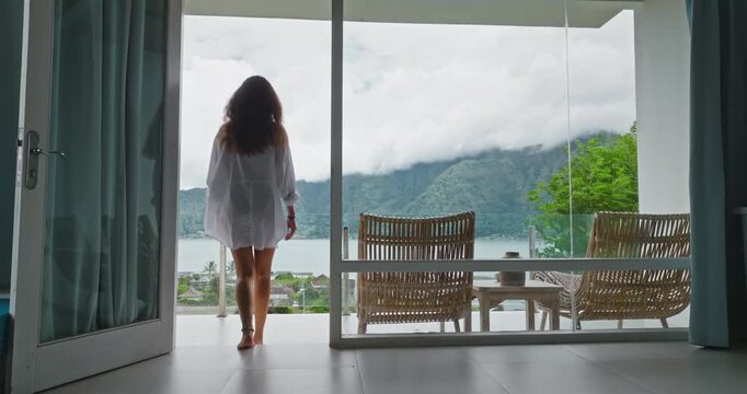 Woman barefoot steps through sliding doors to a modern terrace balcony, breathing fresh morning air, gazing at Lake Batur and green mountains partially shrouded in clouds, serene escape on Bali island