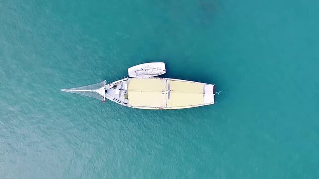 Aerial top-down view of a traditional wooden sailboat with yellow canopy and attached dinghy floating on turquoise ocean water
