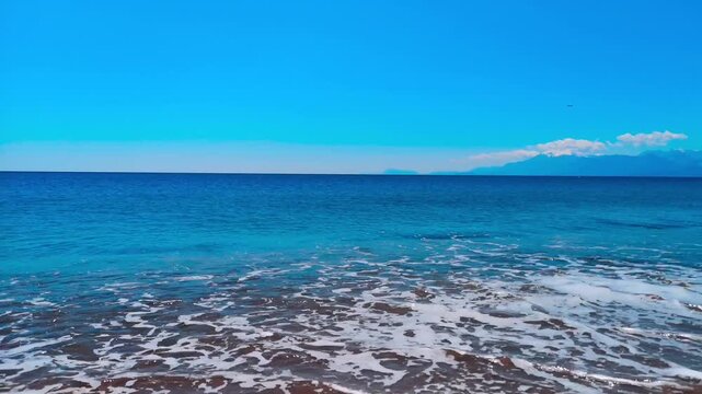 Gentle ocean waves washing ashore on golden sand under bright blue sky with distant mountain range