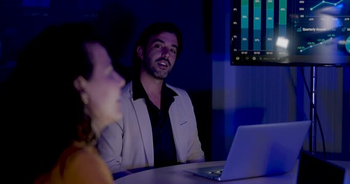 Adult male consultant speaks across meeting table, gestures toward female colleague and references laptop dashboard during business intelligence discussion for data driven strategy and planning.